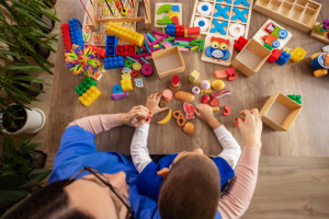 A young boy with autism sits at a table, focused on a colorful sensory toy while a nurse gently guides him.